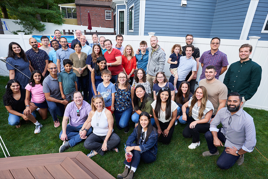 Large group posing in front of white wall and blue house.