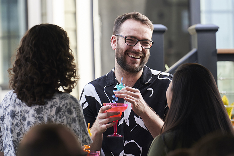 Three people talking at a party. Perosn facing the camera has a drink and other two are only seen from the back.